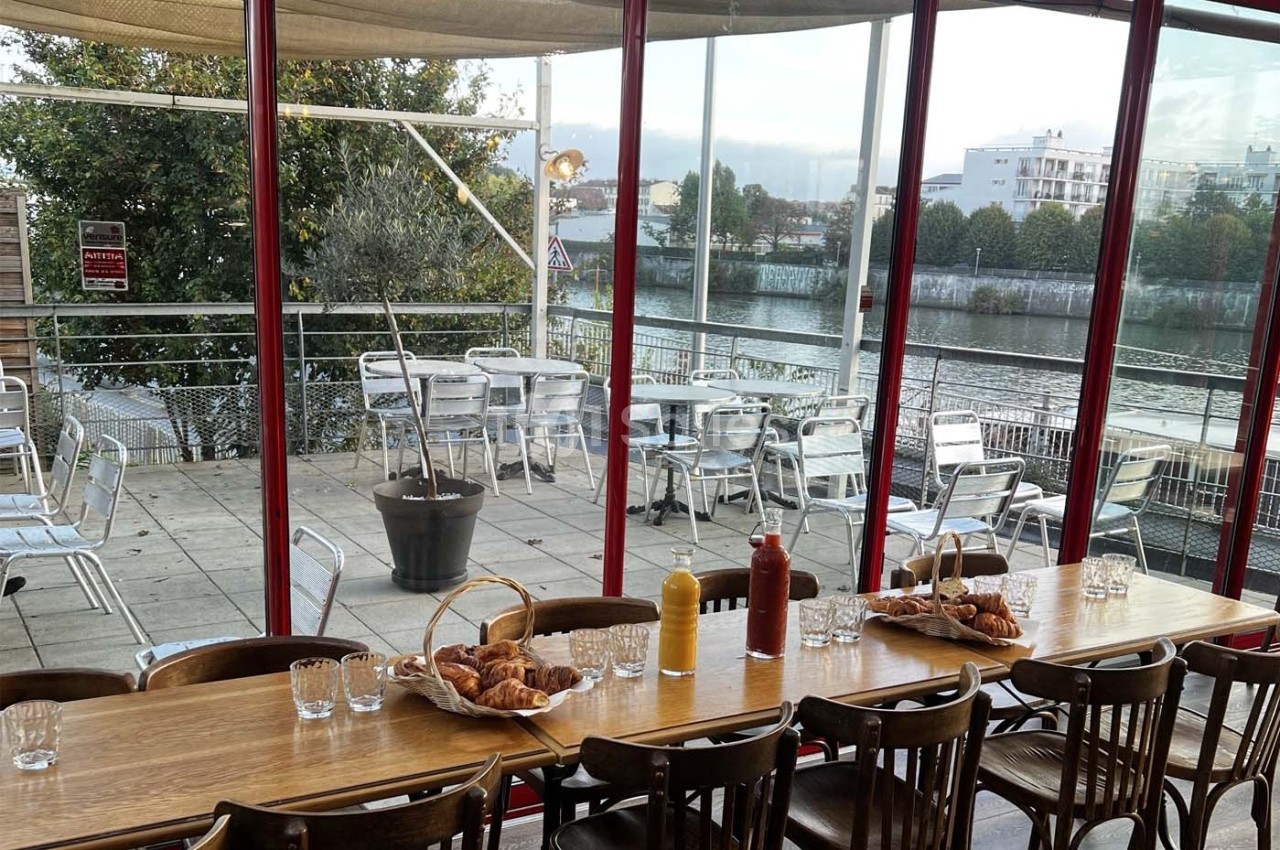 Salle de restaurant avec une grande table en bois, vue sur une terrasse extérieure et un cours d'eau en arrière-plan.