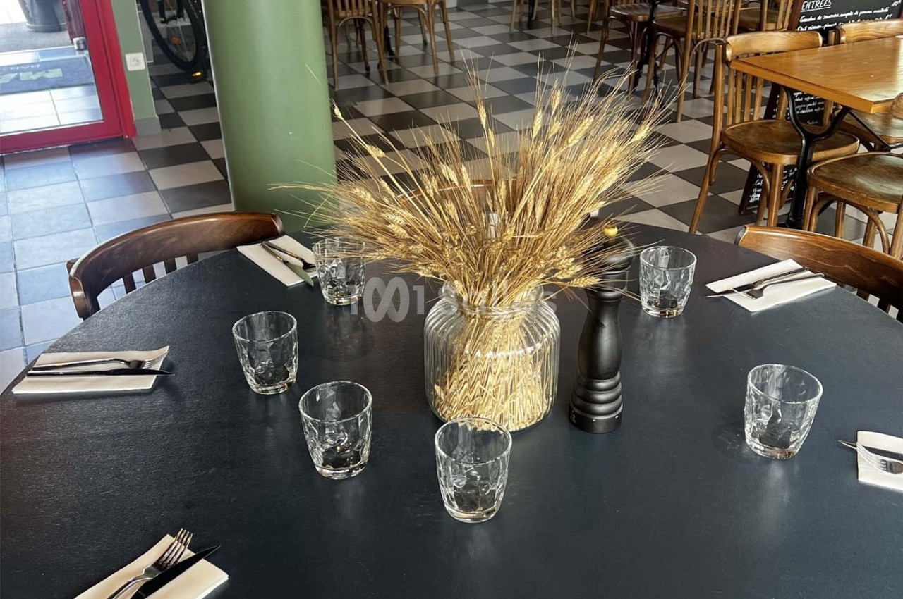 Table ronde dressée avec des verres, couverts et un centre de table en épis de blé dans un restaurant au sol carrelé.