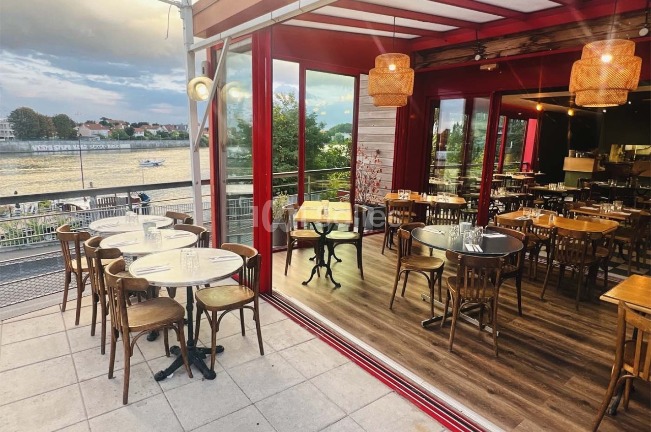 Terrasse et salle intérieure d'un restaurant avec vue sur une rivière, mobilier en bois et ambiance chaleureuse.