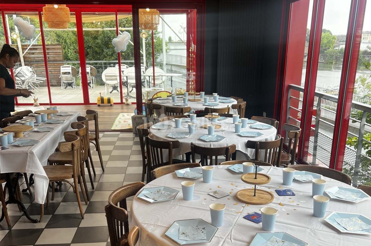 Salle décorée pour un événement avec tables dressées, nappes blanches, vaisselle bleue et vue sur une terrasse extérieure.