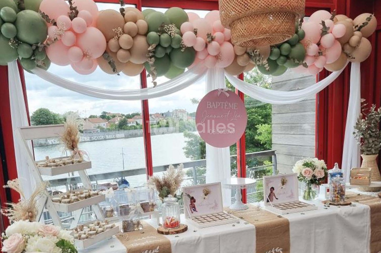 Décoration de baptême avec ballons pastel, table garnie de douceurs et vue sur un paysage extérieur avec rivière.