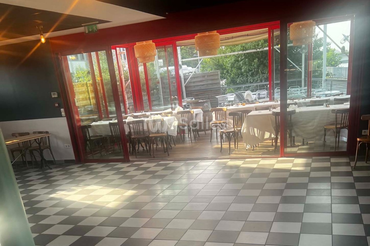 Salle de restaurant lumineuse avec sol en damier, tables dressées derrière une baie vitrée donnant sur une terrasse.