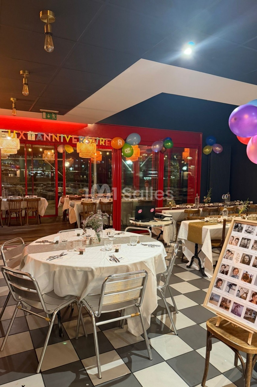 Salle décorée pour une fête avec tables dressées, ballons colorés et un panneau de photos près de l'entrée.