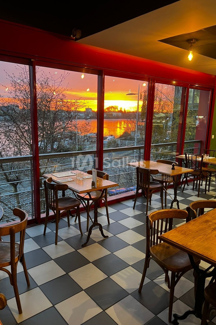 Salle de restaurant avec des tables en bois, grandes baies vitrées et vue sur un coucher de soleil au bord de l'eau.