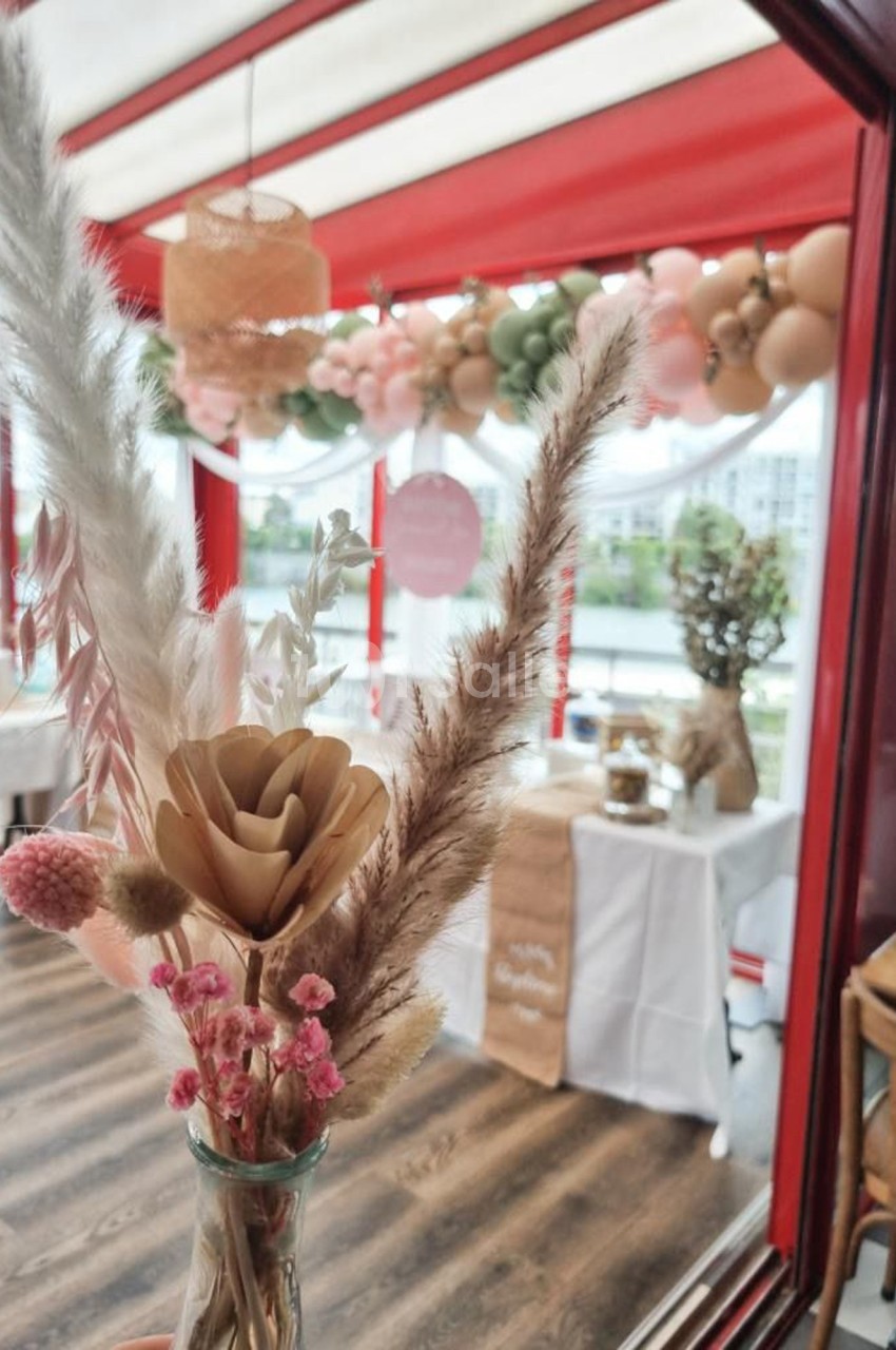 Décoration florale séchée en premier plan, avec une salle festive ornée de ballons et guirlandes en arrière-plan.