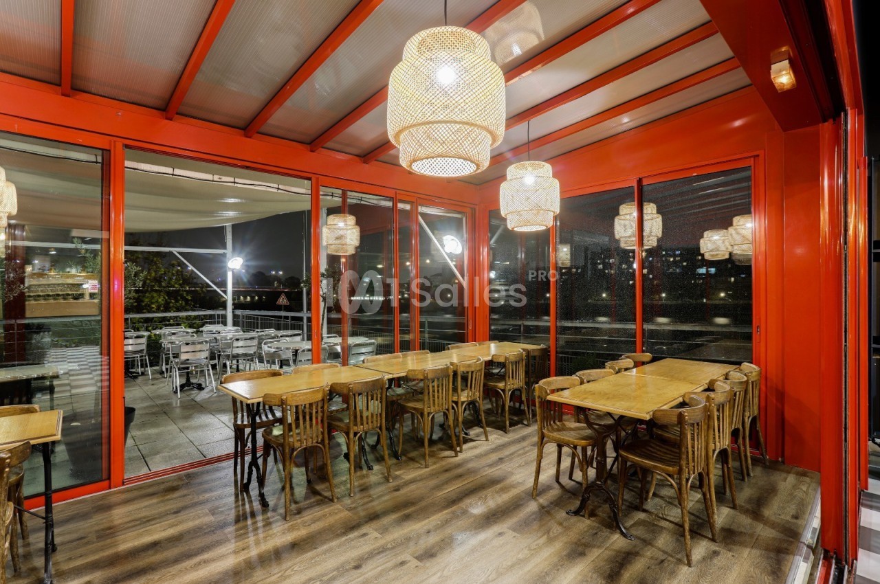 Salle de restaurant avec grandes baies vitrées, mobilier en bois et lampes suspendues, donnant sur une terrasse éclairée.