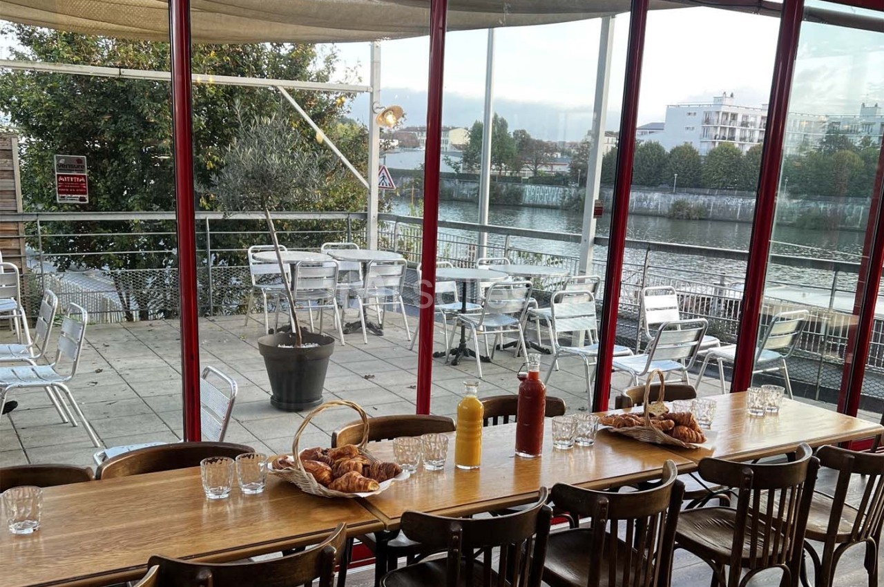 Salle de restaurant avec grande baie vitrée donnant sur une terrasse aménagée et une vue sur un cours d'eau.