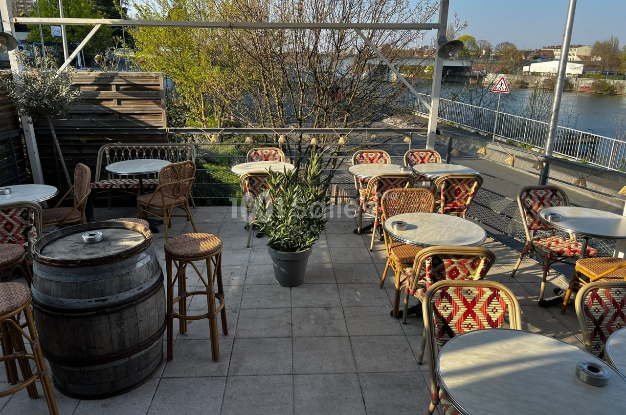 Terrasse extérieure avec tables, chaises en osier, tonneau en bois et vue sur un fleuve bordé de végétation.