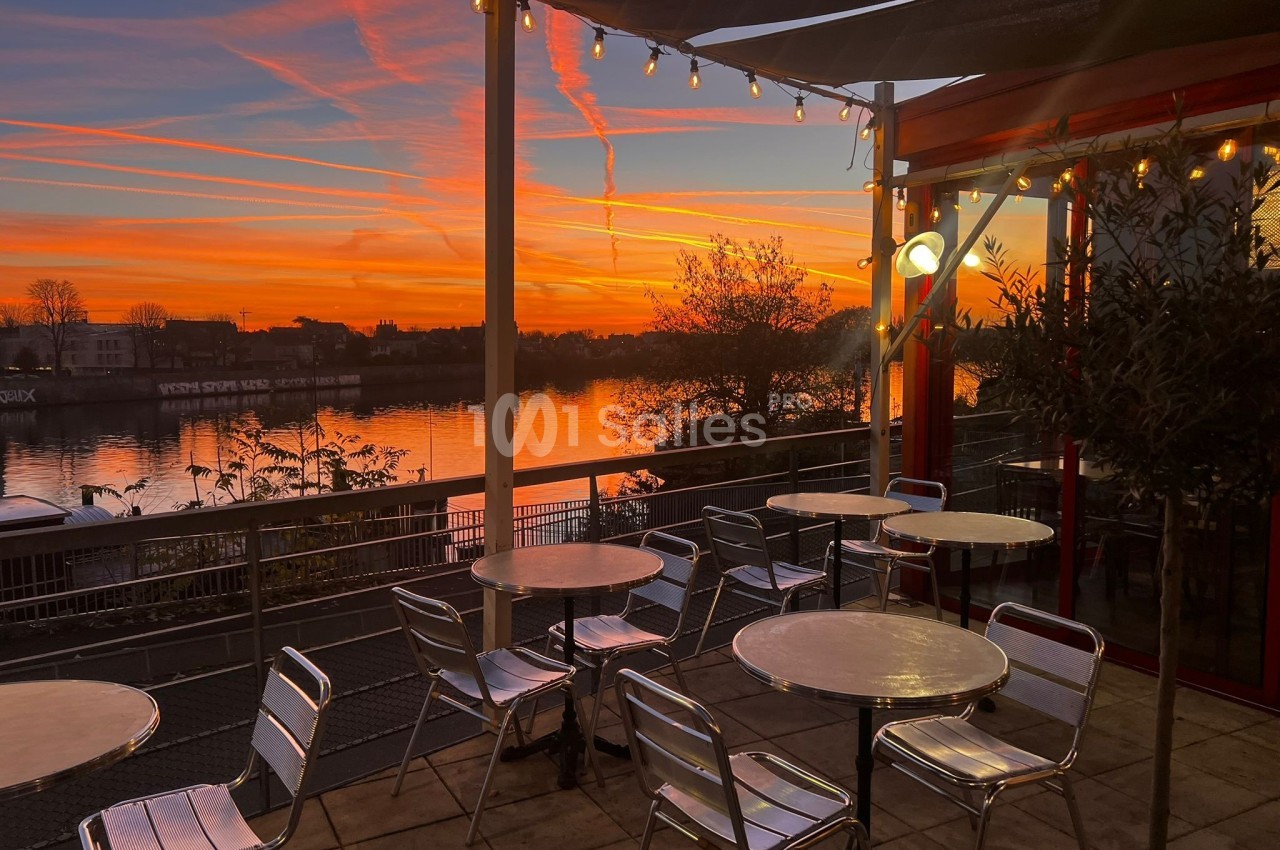 Terrasse avec des tables et chaises métalliques, vue sur un fleuve au coucher du soleil avec un ciel orangé.