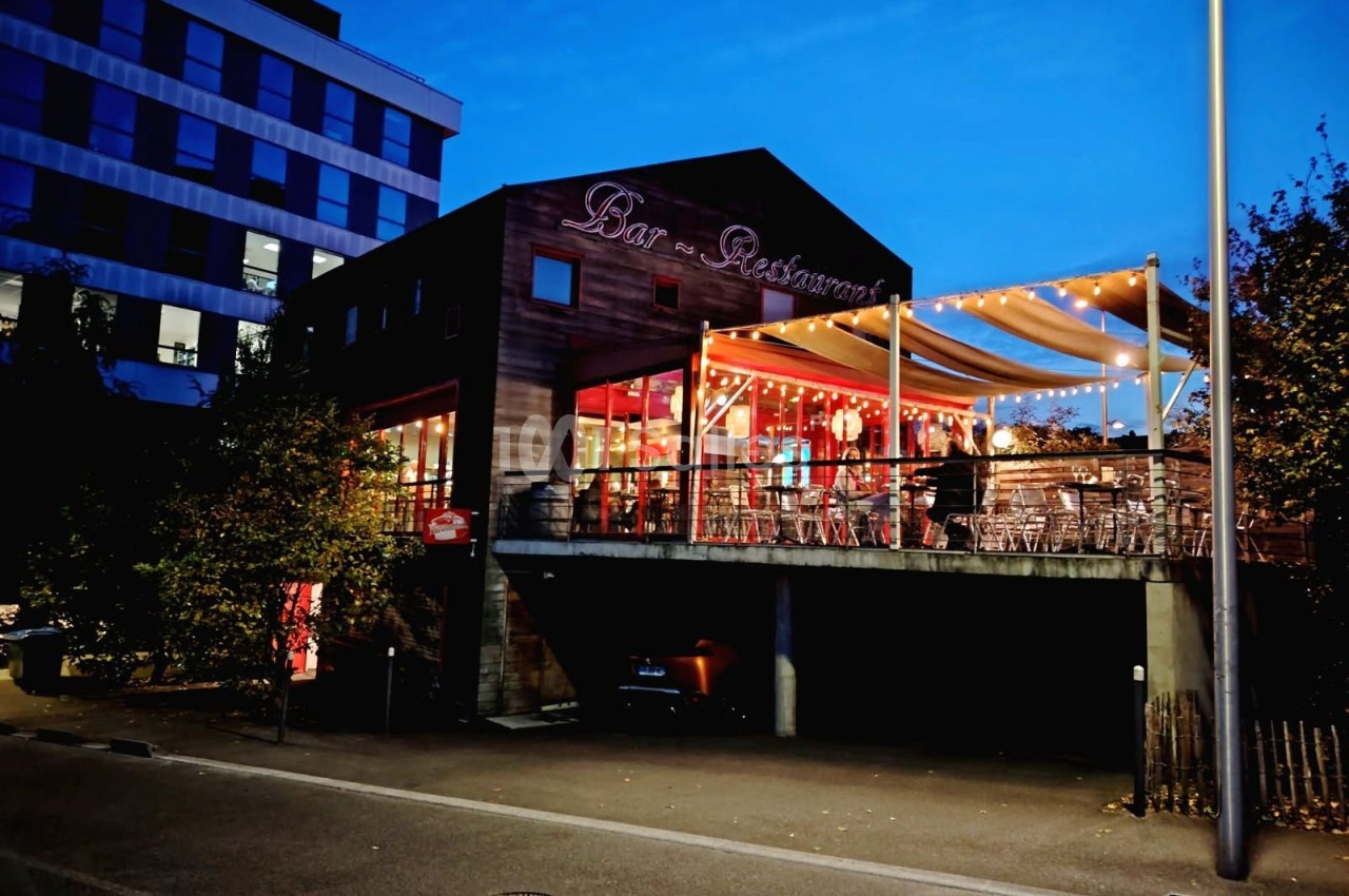 Terrasse d'un bar-restaurant éclairée en soirée, avec des tables et chaises sous une pergola lumineuse.
