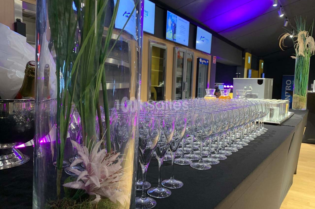 Rangée de verres à vin alignés sur une table noire, avec des vases décoratifs et un éclairage violet en arrière-plan.