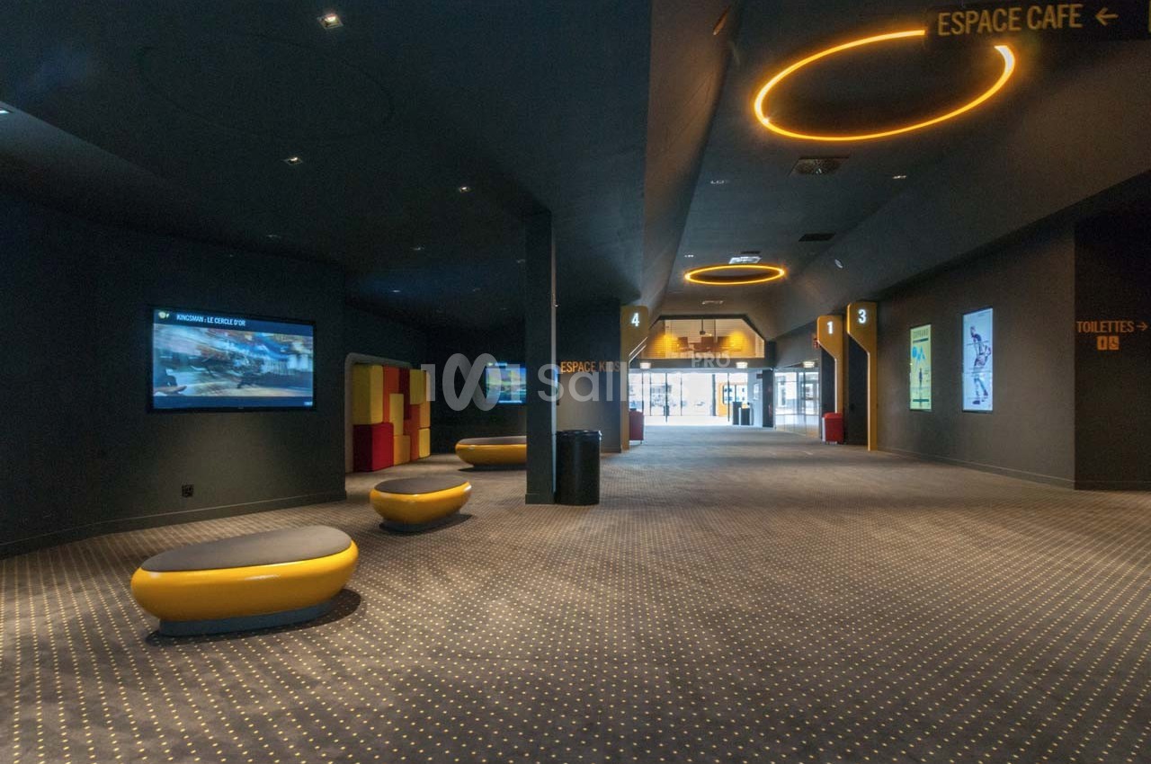 Couloir d'un cinéma avec sièges arrondis, écrans muraux et signalisation vers l'espace café et les toilettes.