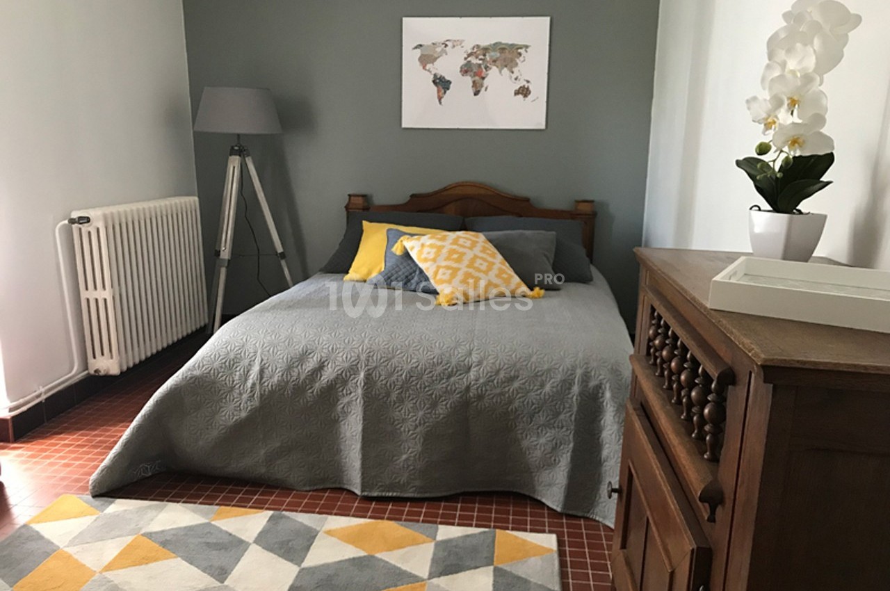 Chambre avec un lit double, coussins jaunes et gris, mur vert, lampe sur pied, carte du monde et orchidée blanche.