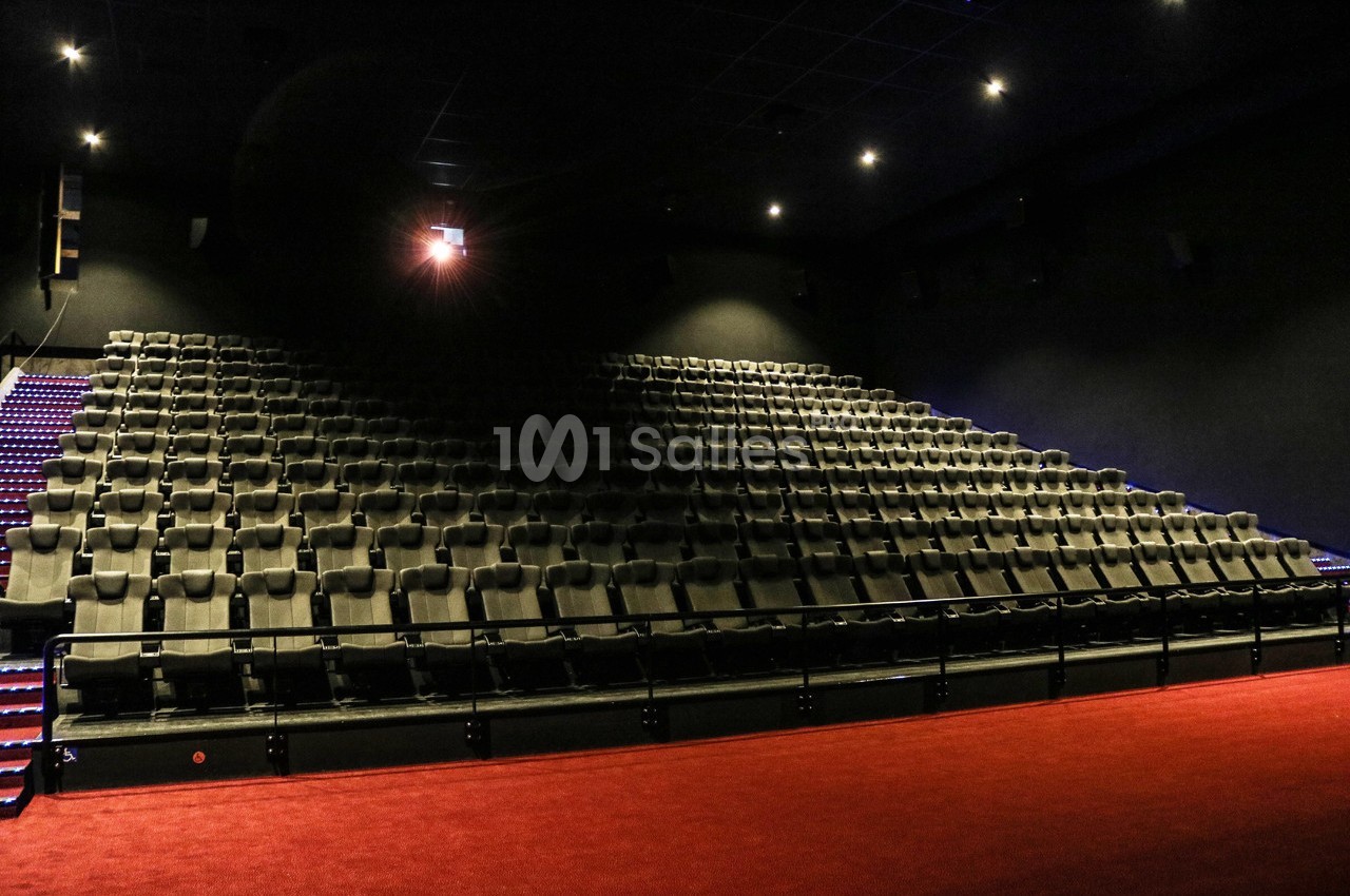 Salle de cinéma vide avec rangées de sièges noirs, éclairée par une faible lumière provenant du projecteur.