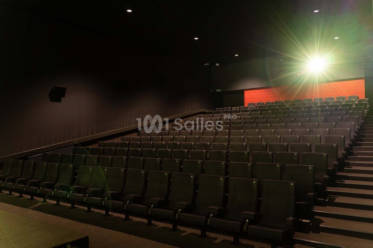 Salle de cinéma vide avec des rangées de sièges noirs et un écran illuminé en arrière-plan.