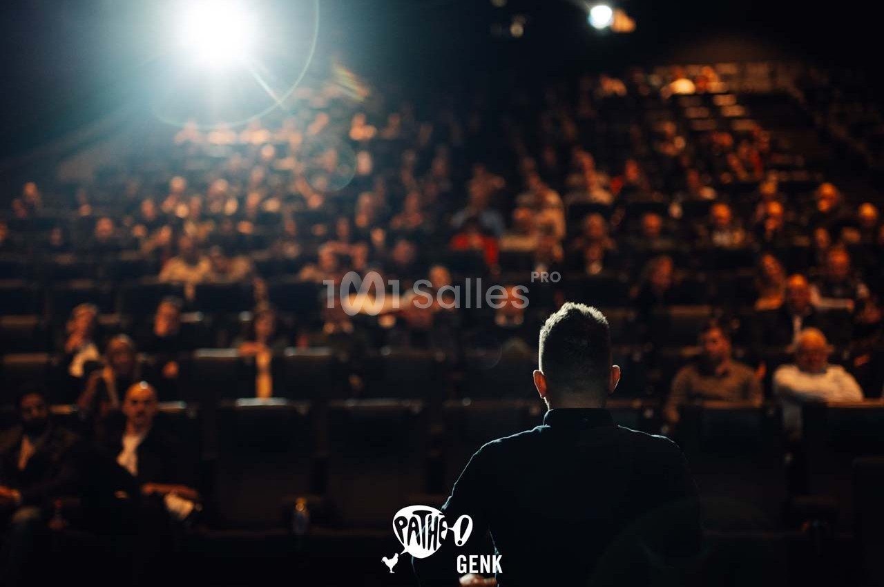 Une personne s'adresse à un public assis dans une salle de spectacle sombre, éclairée par des projecteurs.