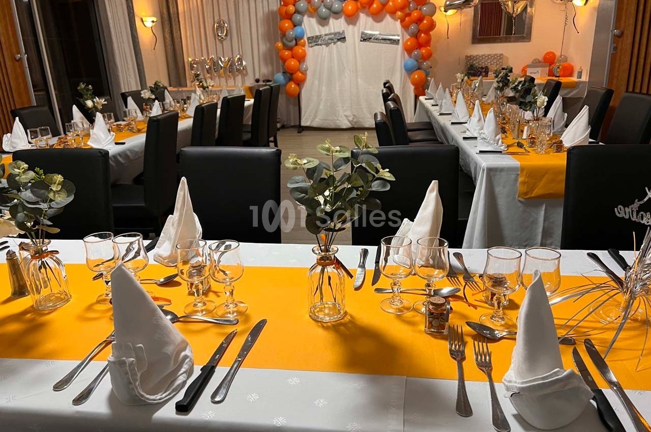 Salle décorée pour une réception avec tables dressées, chemins de table orange et ballons en arrière-plan.