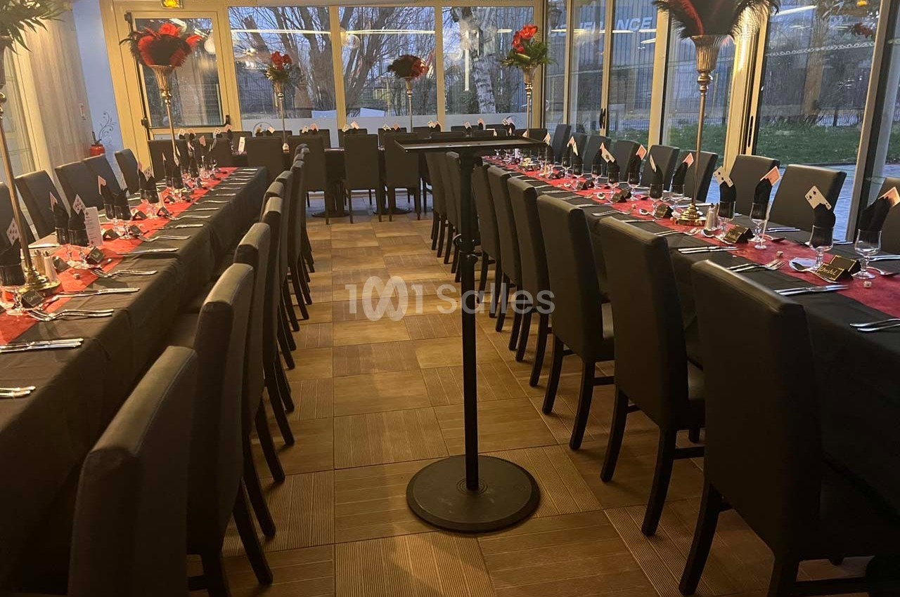 Salle de réception avec deux longues tables dressées, nappes rouges, chaises noires et pupitre central.