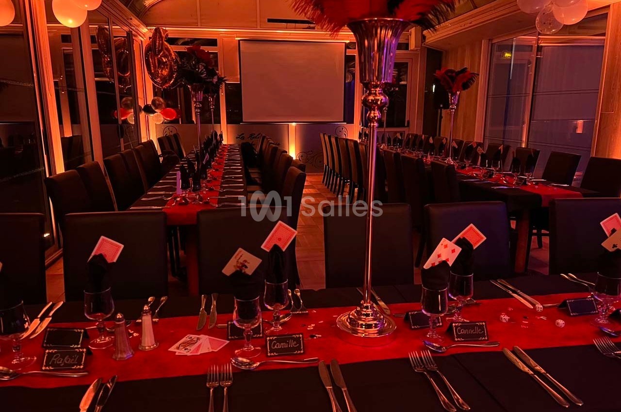 Salle décorée pour un événement, avec tables alignées, nappes rouges et noires, cartes à jouer et plumes en centre de table.