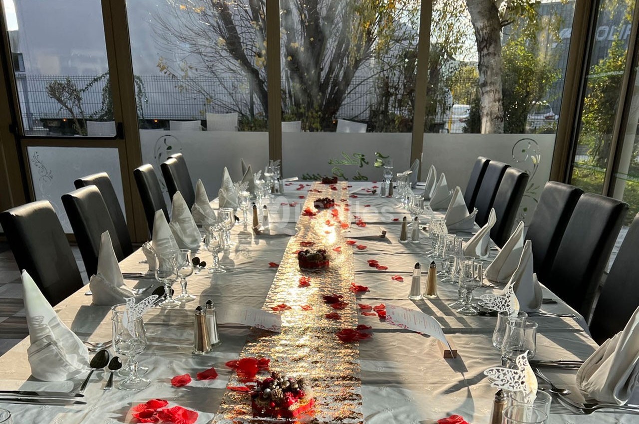 Table élégamment dressée avec nappes blanches, serviettes pliées, pétales rouges et lumière naturelle en arrière-plan.