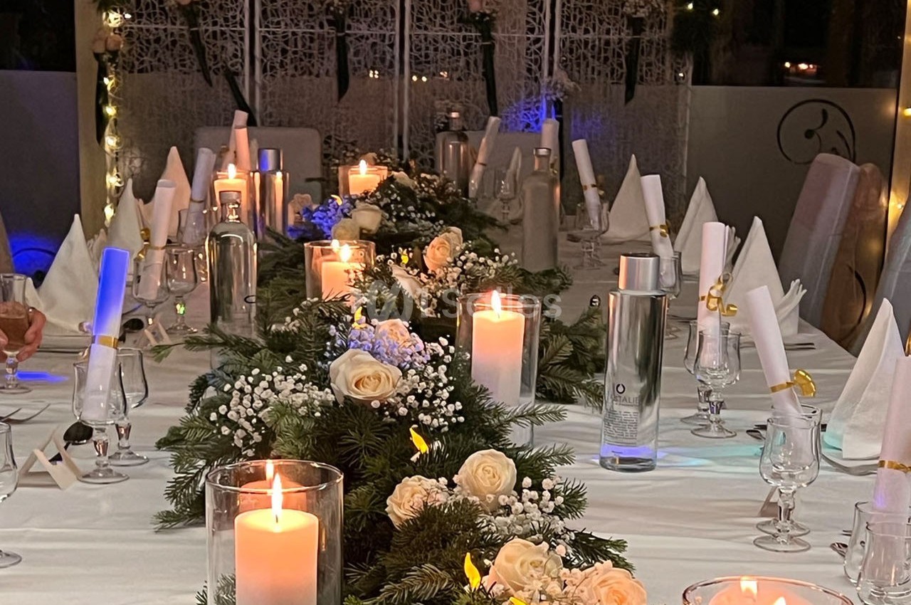 Table décorée pour un dîner festif avec bougies allumées, fleurs blanches et serviettes pliées sur une nappe blanche.
