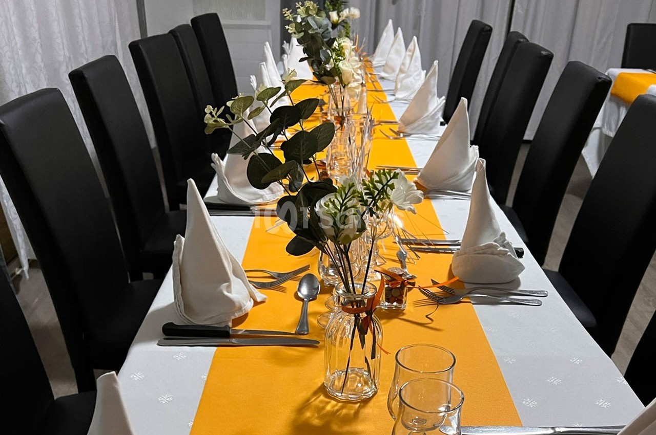 Table dressée avec nappes blanches, chemins de table orange, serviettes pliées, couverts et décorations florales.
