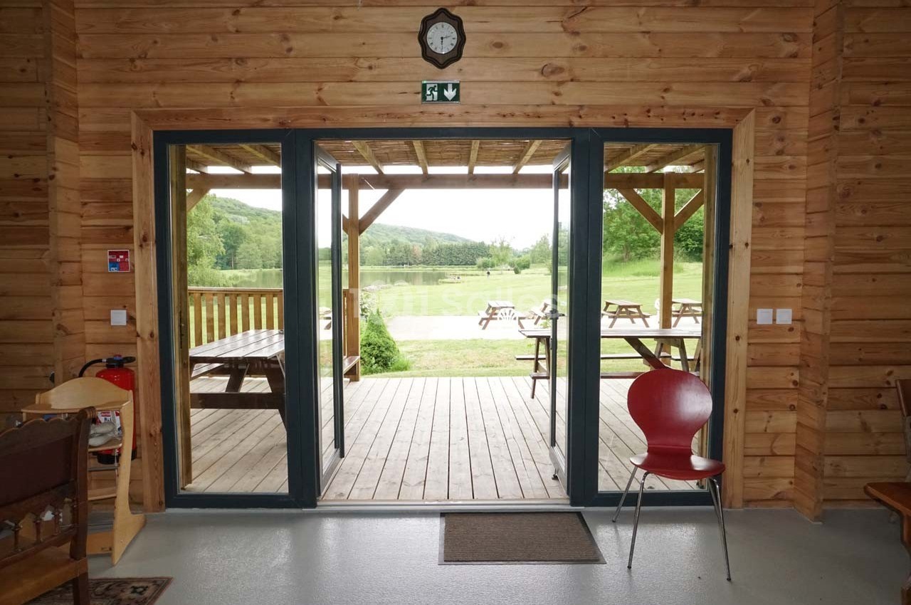 Vue depuis une pièce en bois sur une terrasse donnant sur un espace extérieur avec tables de pique-nique et paysage…