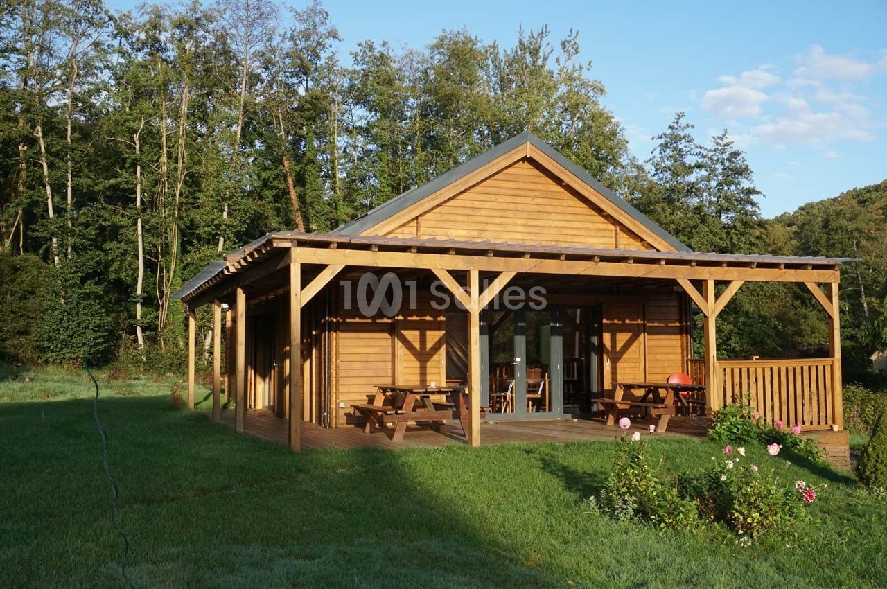 Chalet en bois avec terrasse couverte, entouré de pelouse et d'arbres, sous un ciel partiellement dégagé.