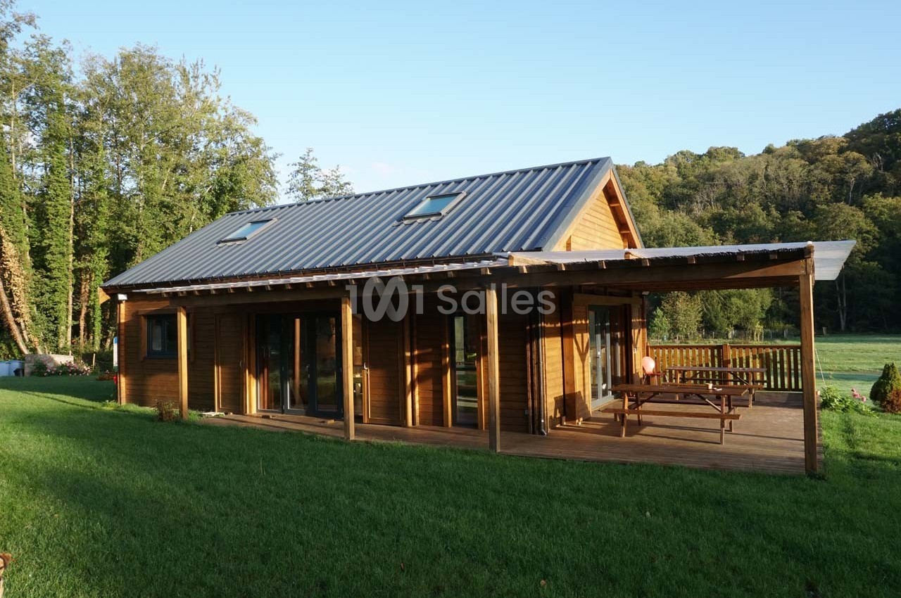 Maison en bois avec toit en métal, terrasse couverte et table de pique-nique, entourée de pelouse et d'arbres.