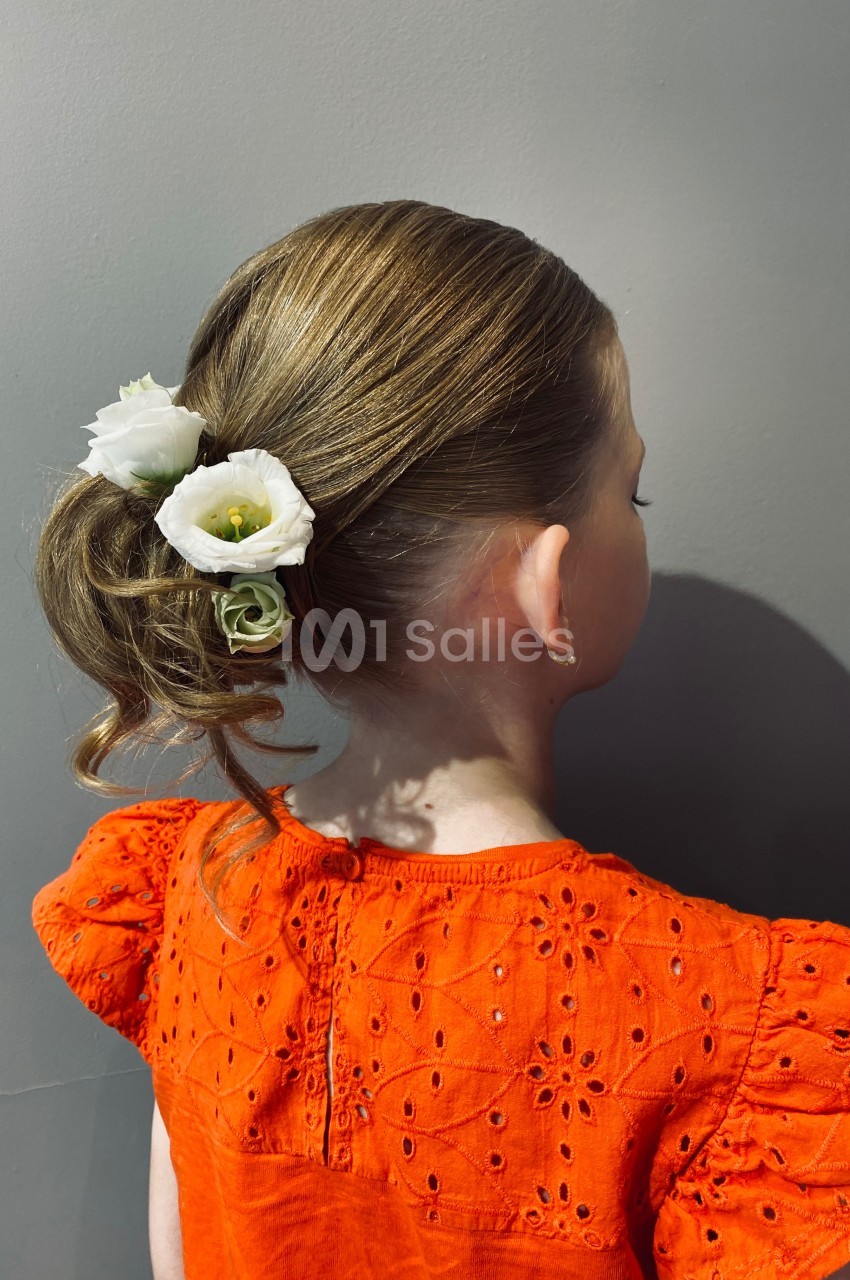 Une fillette de dos avec une coiffure relevée ornée de fleurs blanches, portant une robe orange à motifs brodés.