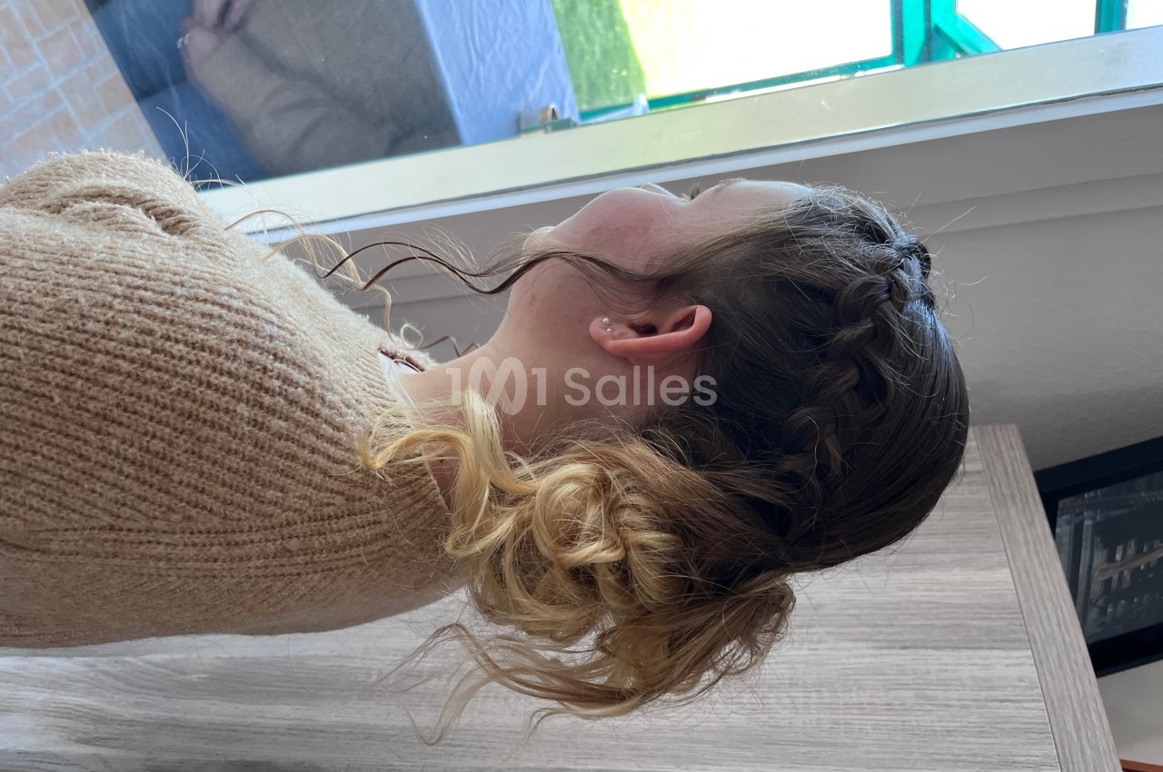 Femme de dos avec des cheveux blonds coiffés en tresse et chignon, portant un pull beige près d'une fenêtre.
