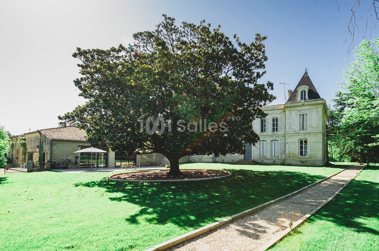 Grand arbre au centre d'un jardin verdoyant, avec une maison en pierre et une allée gravillonnée en arrière-plan.