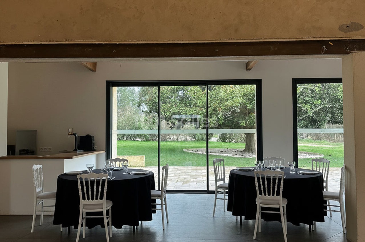 Salle lumineuse avec tables rondes dressées, nappes noires, chaises blanches et vue sur un jardin verdoyant par de grandes…