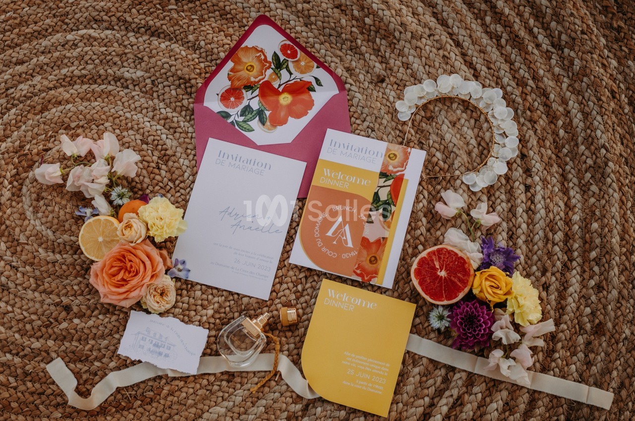 Ensemble d'invitations colorées et décoratives posées sur un tapis en fibres naturelles, entourées de fleurs et d'agrumes.