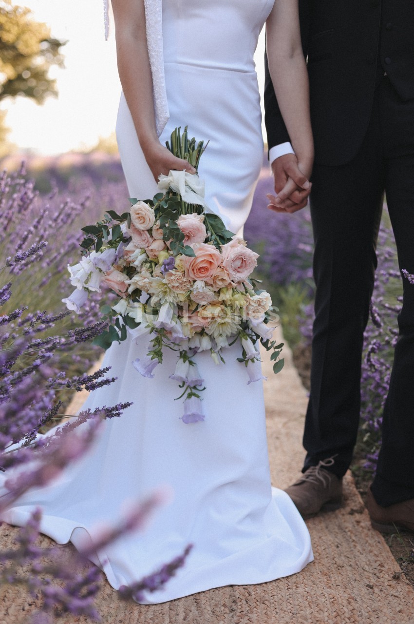 Un couple en tenue de mariage se tient la main dans un champ de lavande, avec un bouquet de fleurs pastel.