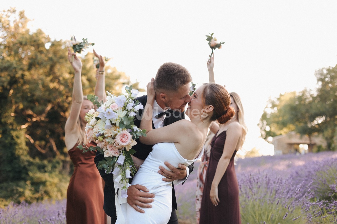 Un couple de mariés s'embrasse dans un champ de lavande, entouré de demoiselles d'honneur levant des bouquets.