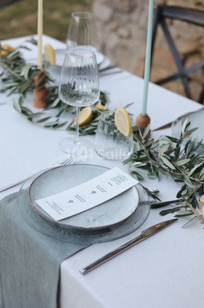 Table dressée avec assiette, menu, verres, bougies et décoration végétale incluant des branches d'olivier et des tranches de…