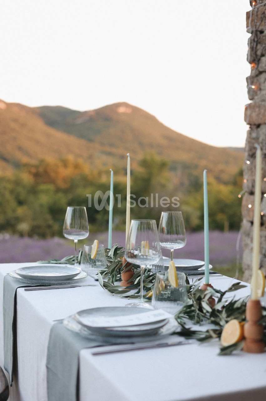 Table dressée en extérieur avec nappes blanches, bougies, verres à pied et décorations végétales devant un paysage…
