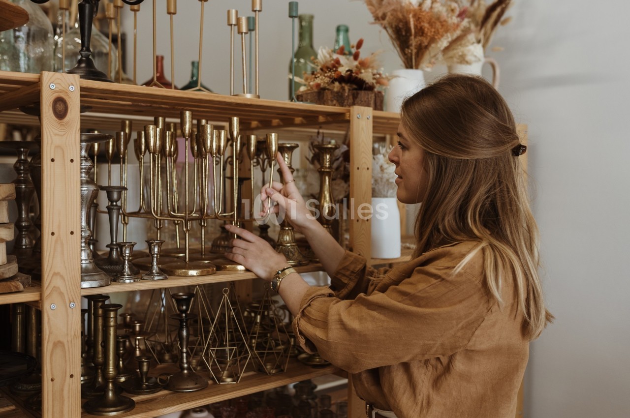 Une femme examine des chandeliers en métal doré sur une étagère en bois dans un magasin ou un atelier.