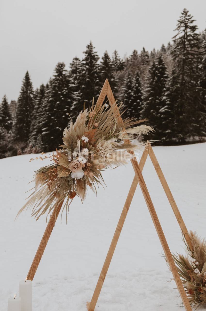 Structure en bois triangulaire décorée de fleurs séchées et de pampas, placée dans un paysage enneigé avec des sapins.