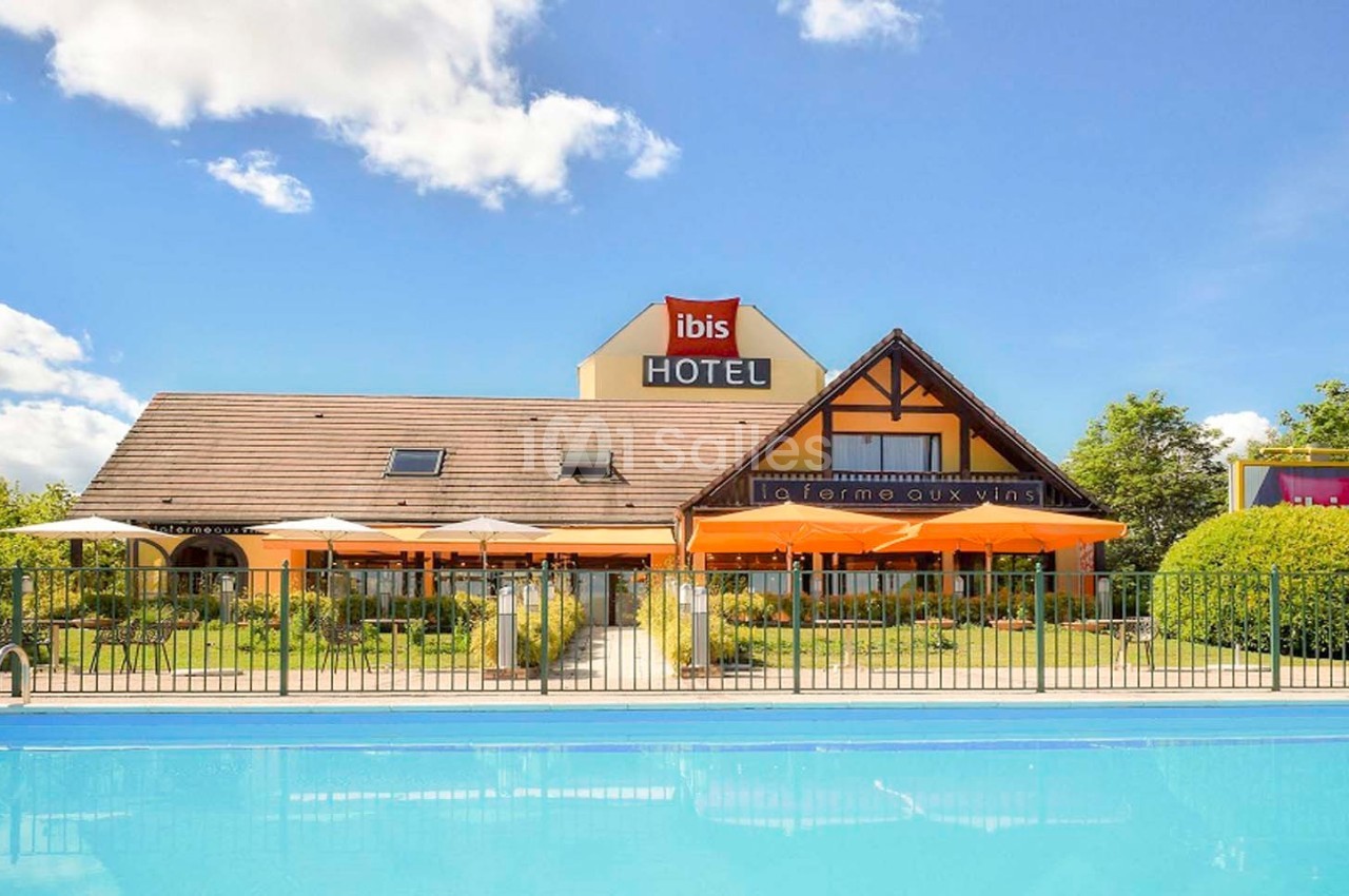 Façade d'un hôtel avec terrasse, parasols et jardin, vue depuis une piscine entourée d'une barrière.