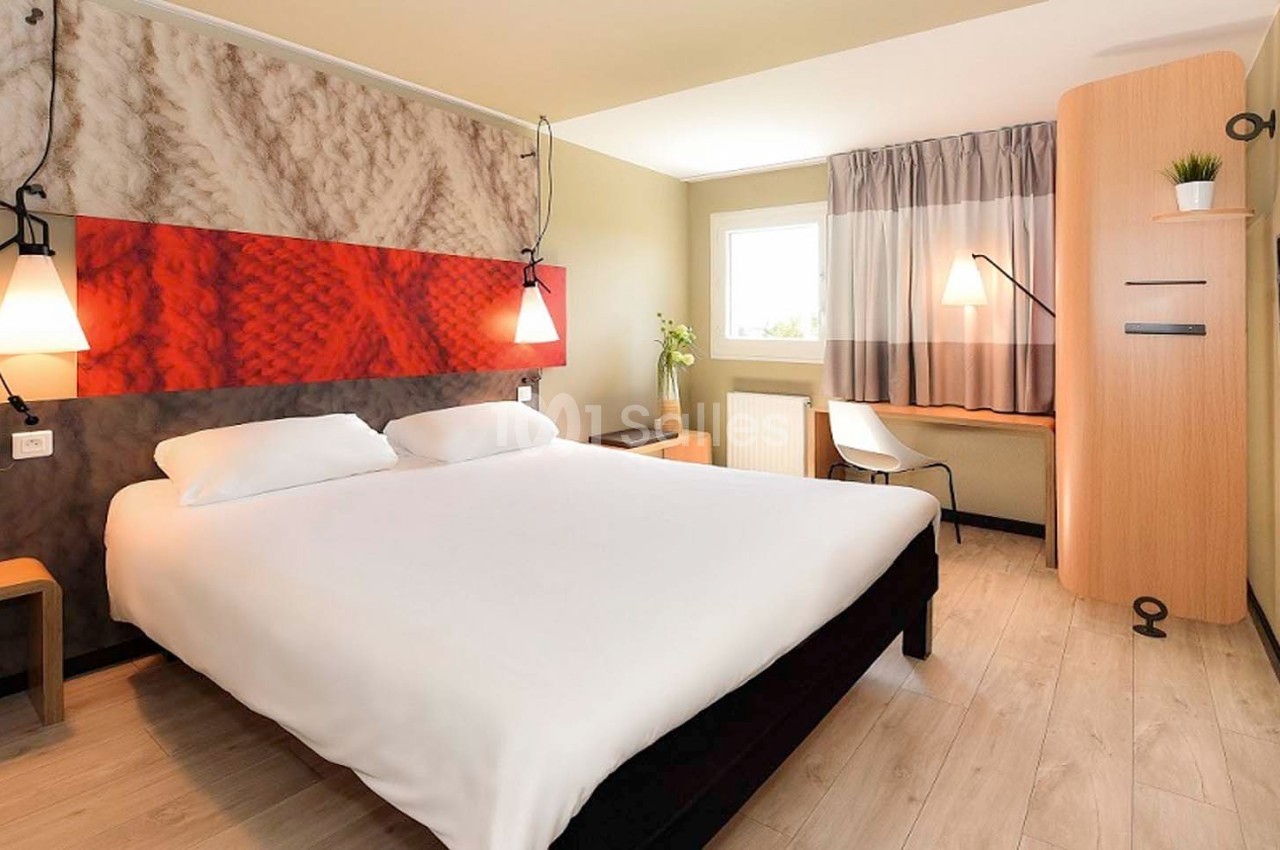Chambre d'hôtel avec lit double, bureau en bois, lampe suspendue, fenêtre et décoration murale rouge et beige.