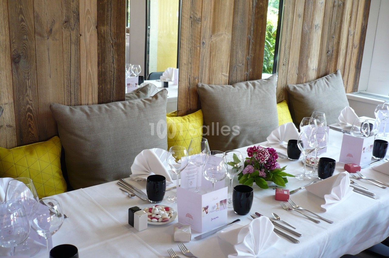 Table dressée avec nappes blanches, serviettes pliées, verres, fleurs et coussins jaunes sur une banquette en bois.
