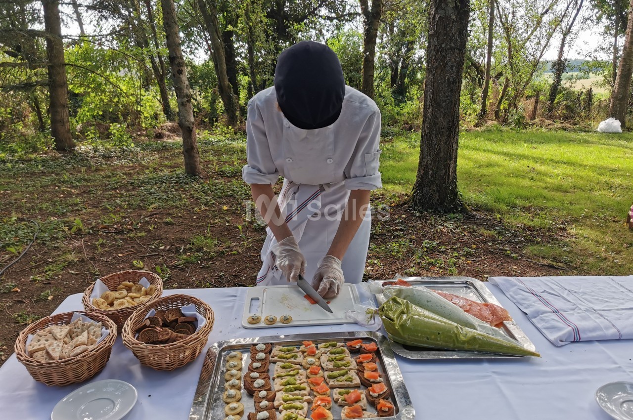 Un chef prépare des amuse-bouches sur une table dressée en extérieur, entourée d'arbres et de verdure.