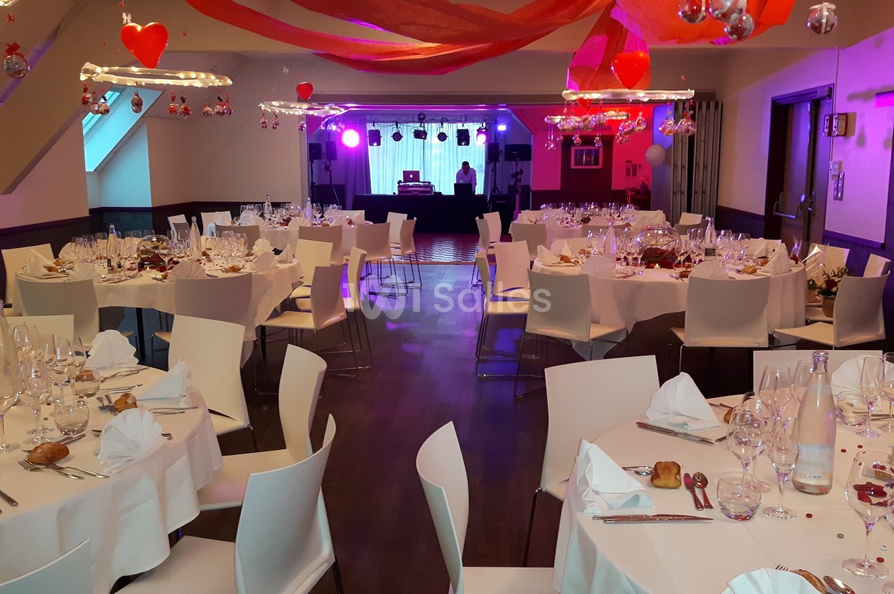 Salle décorée pour un événement avec des tables dressées, des cœurs suspendus et une scène avec éclairage coloré.
