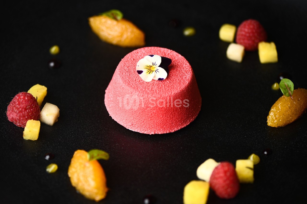Dessert rose texturé garni d'une fleur comestible, entouré de fruits frais et de petites touches de sauce sur une assiette…