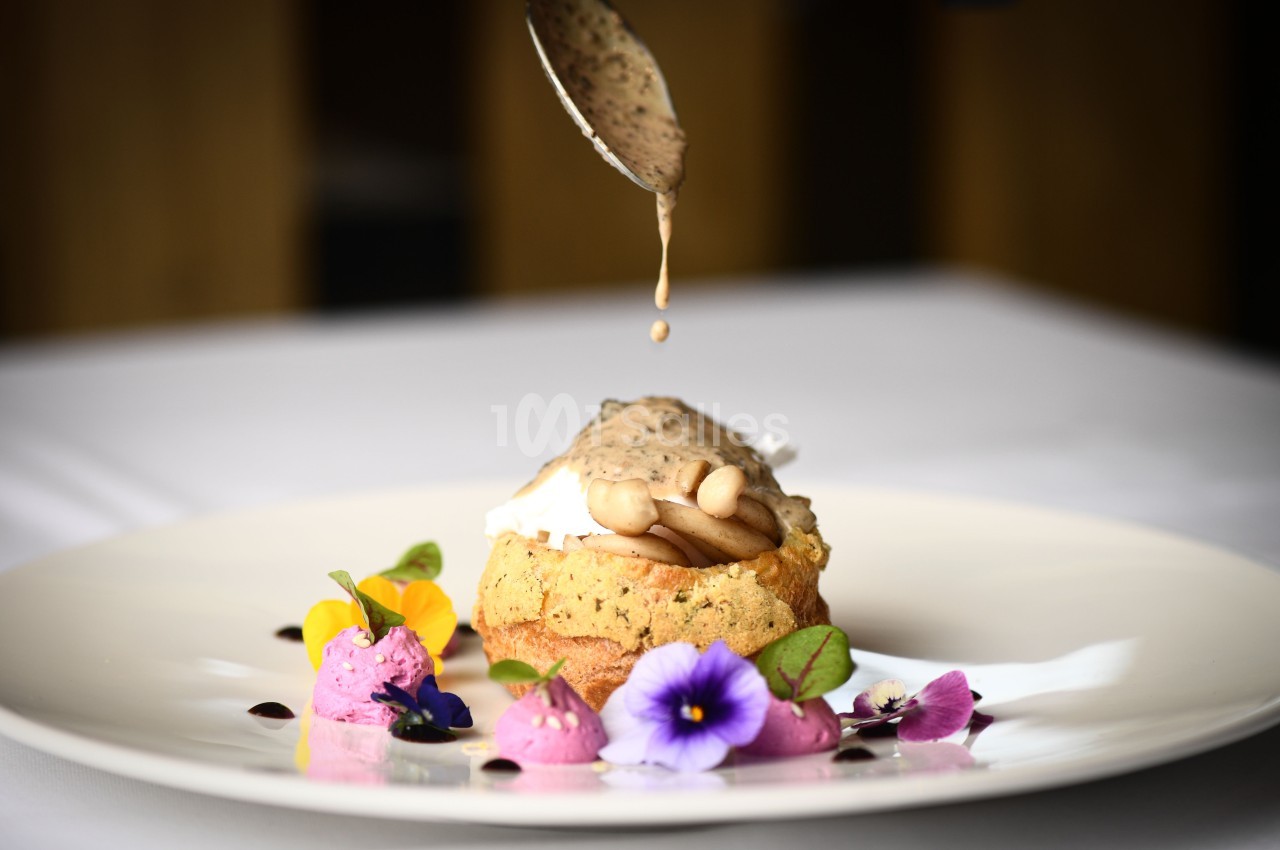 Un plat raffiné avec une sauce versée sur un soufflé garni de champignons, accompagné de fleurs comestibles.