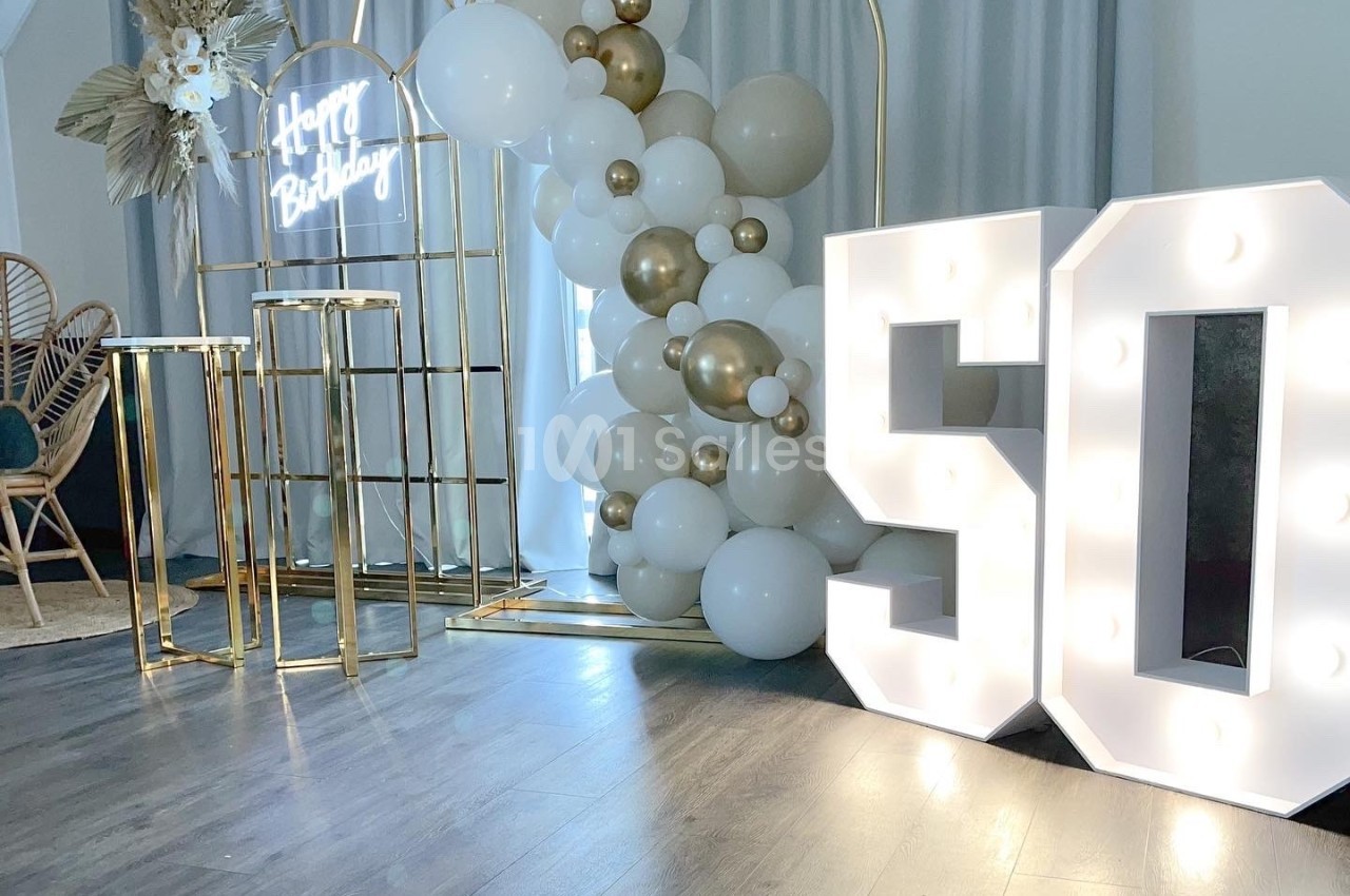 Décoration d'anniversaire avec ballons blancs et dorés, arche métallique, et chiffres lumineux ’50’ sur un sol en bois.