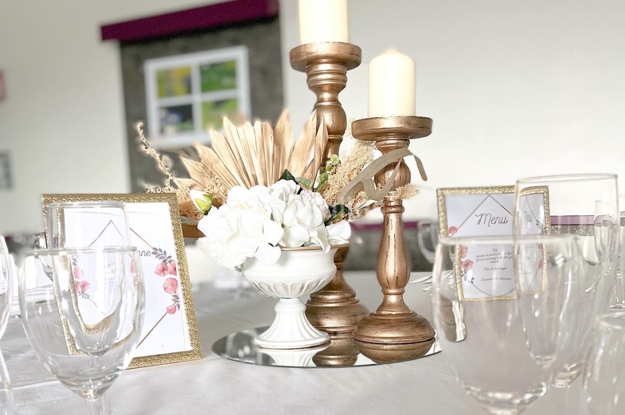 Centre de table avec chandeliers dorés, bougies, fleurs blanches et cadres décoratifs sur une nappe blanche.