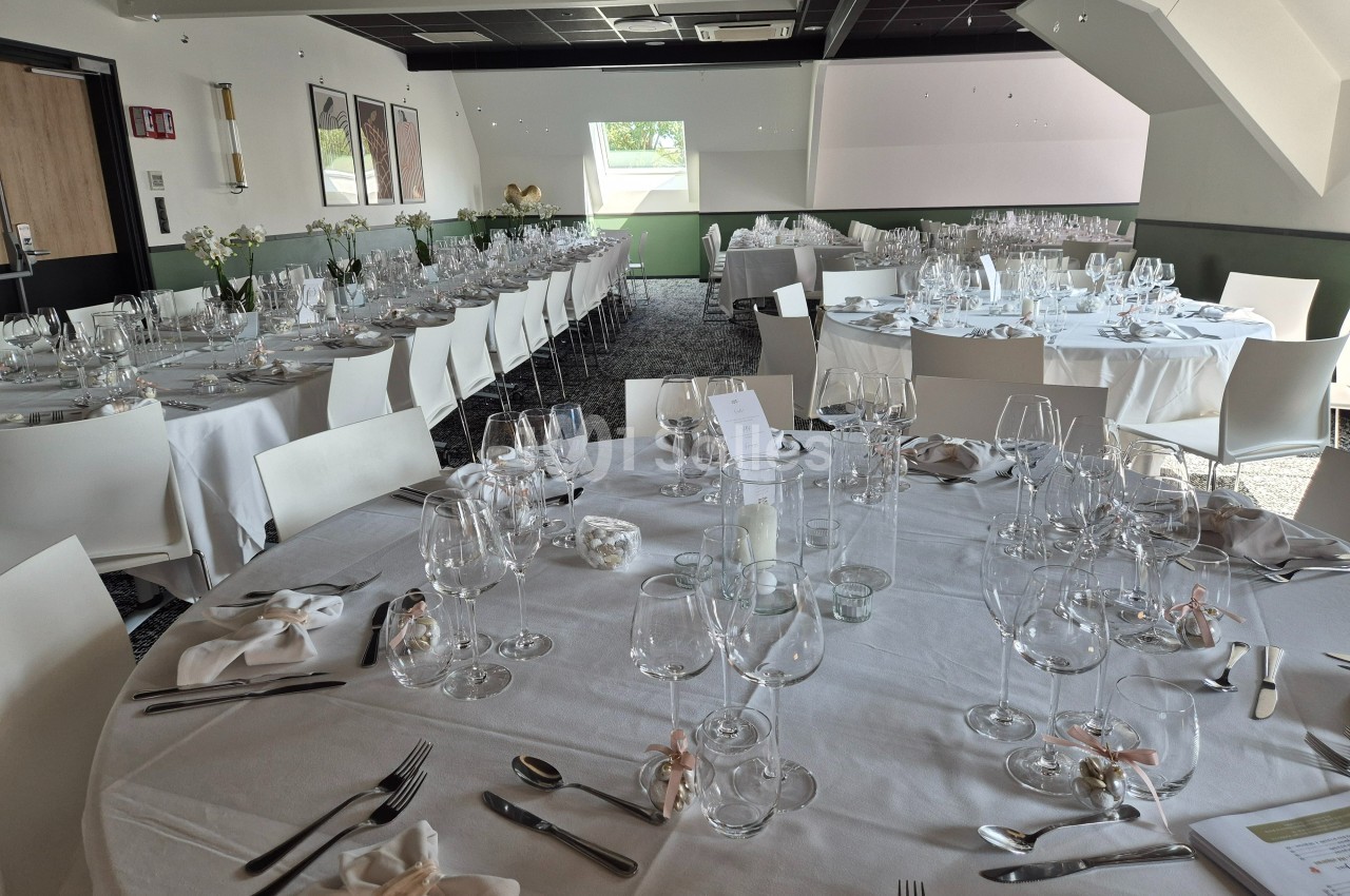 Salle de réception lumineuse avec des tables rondes dressées pour un repas, ornées de verres et décorations élégantes.